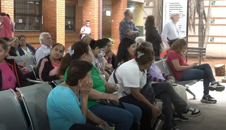 Un grupo de adultos en sala de espera, algunos con mascarillas, expresando tristeza y tranquilidad en un ambiente hospitalario.
