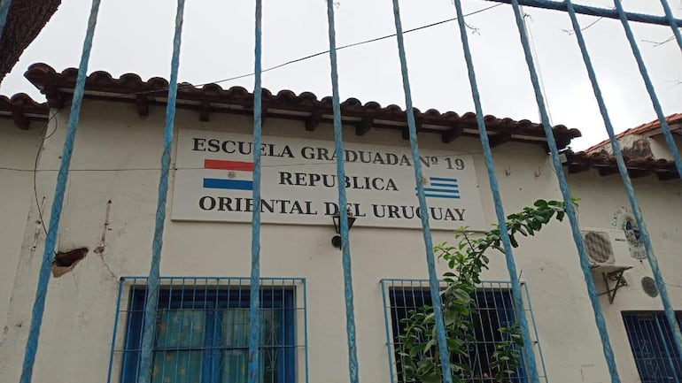 Fachada de la escuela República del Uruguay con reja azul y carteles visibles, rodeada de árboles y plantas verdes.