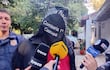 Persona con chaqueta negra y capuchón, rodeada de reporteros con micrófonos en un entorno al aire libre.