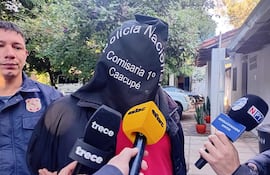 Persona con chaqueta negra y capuchón, rodeada de reporteros con micrófonos en un entorno al aire libre.