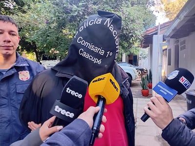 Persona con chaqueta negra y capuchón, rodeada de reporteros con micrófonos en un entorno al aire libre.