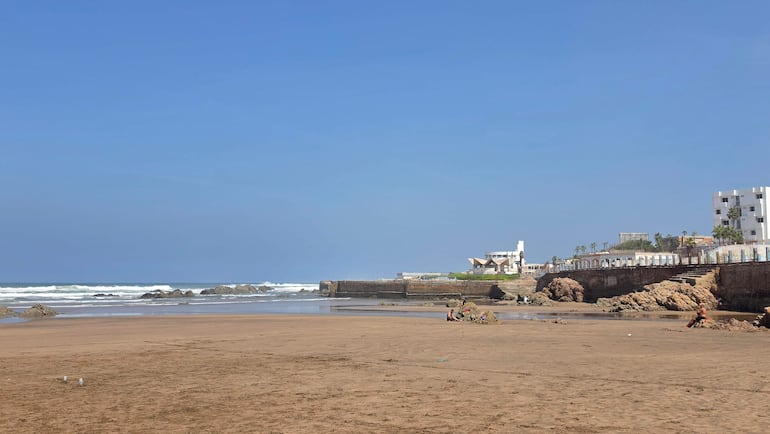 Personas dispersas en la arena de la playa, un grupo interactúa, con edificios modernos y tradicionales al fondo.