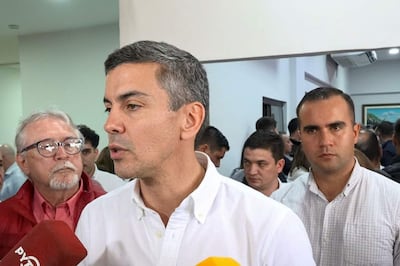 Santiago Peña en camisa blanca frente a micrófono rojo, rodeado de personas en un ambiente formal, algunos observando atentamente.
