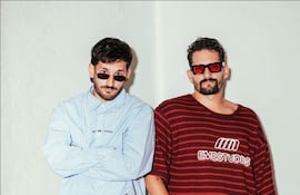 Mau y Ricky de pie, con Mau en camisa de cuadros azul y gafas de sol, y Ricky en camiseta de rayas rojas y negras.