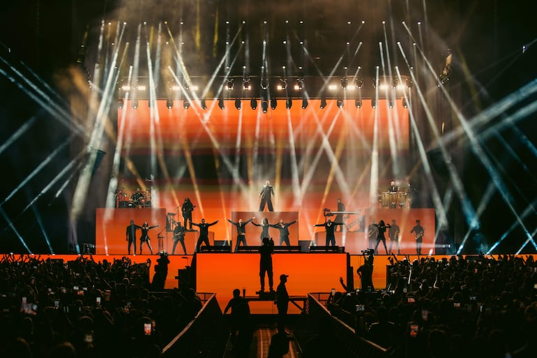 Ricky Martin en el centro del escenario, brazos extendidos, rodeado de músicos en atuendos oscuros, luces intensas y audiencia grabando con celulares.