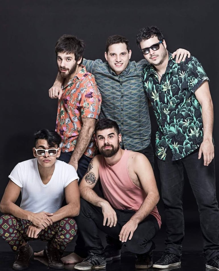 Cinco personas posan en un fondo oscuro, mostrando sonrisas y diversas vestimentas coloridas, reflejando alegría y diversión.