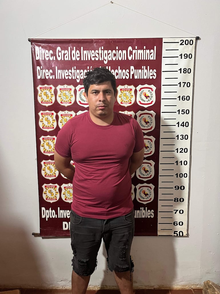 Hombre de pie con camiseta roja y pantalones cortos, serio, frente a un cartel de la investigación criminal en ambiente interior.