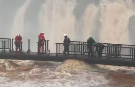 Cinco operarios, dos con trajes rojos y cascos blancos, ajustan equipos en un entorno de cataratas con agua marrón intensa.