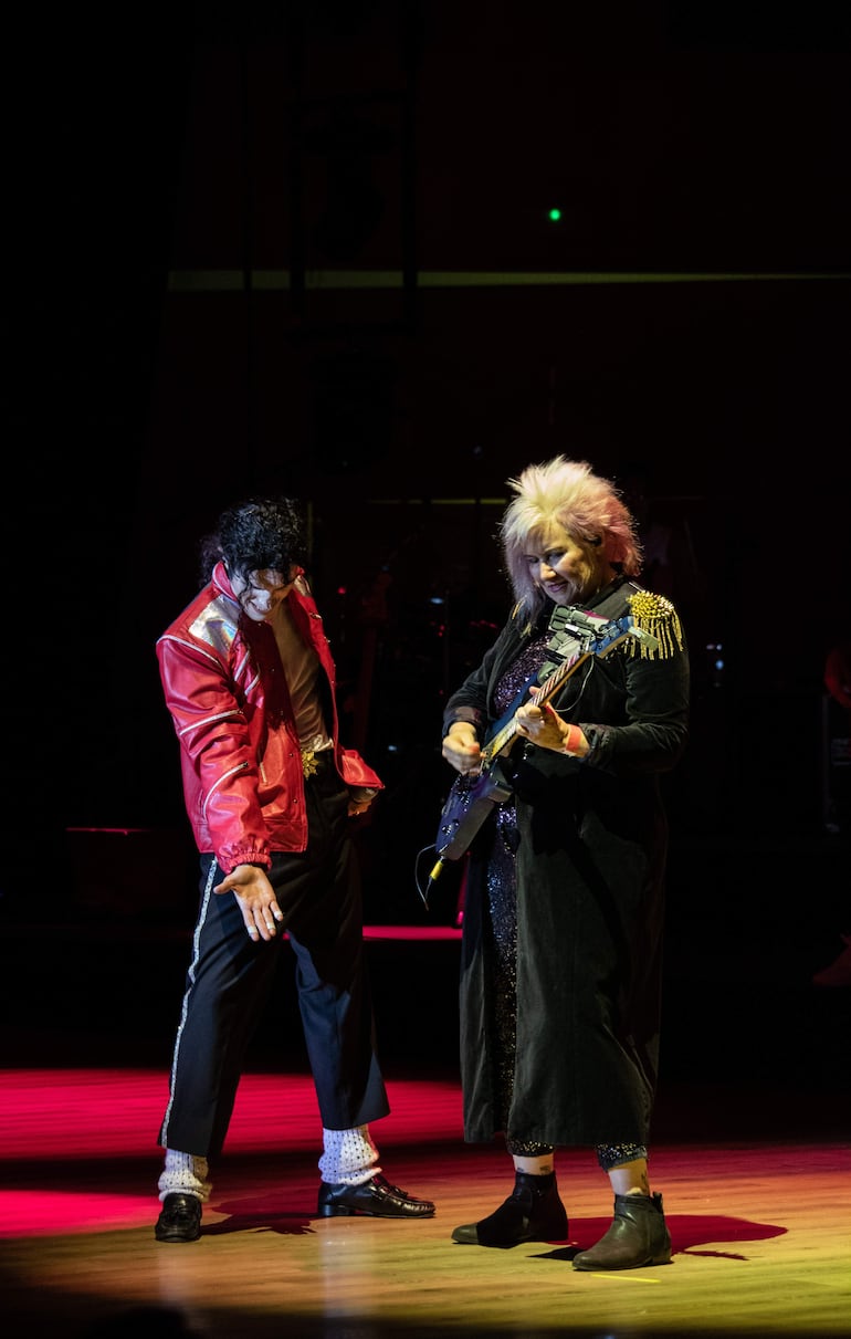 Lenny Jay con chaqueta roja y pantalones oscuros, y Jennifer Batten en abrigo negro, tocando guitarra eléctrica iluminados por luces rojas y amarillas.