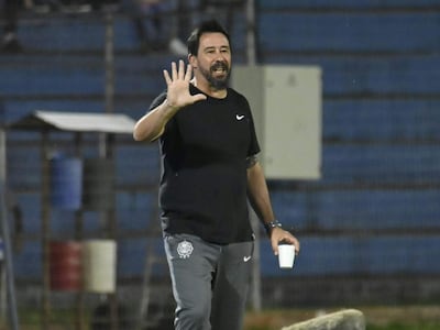 Pablo Sánchez Vitamina en camisa negra y pantalones grises, gesticulando mientras sostiene un vaso, rodeado por público en gradas.