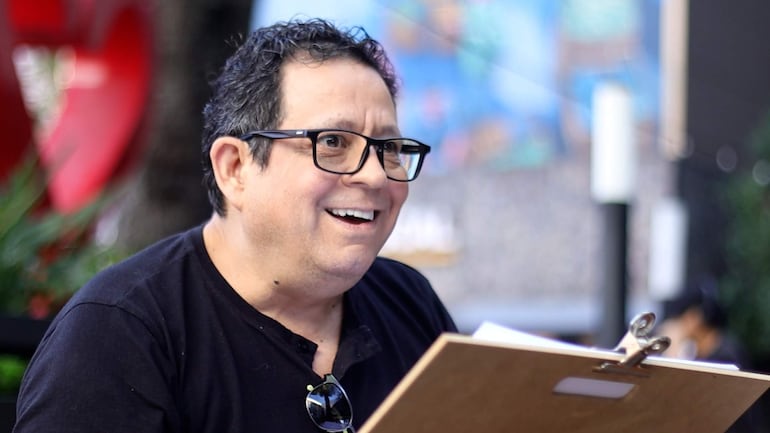 Hombre de cabello oscuro y rizado, con gafas y camiseta negra, sonríe mientras sostiene un portapapeles en un entorno luminoso.