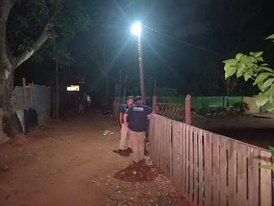 Dos policías en uniforme observan un área en una calle oscura con vegetación y una construcción iluminada al fondo.