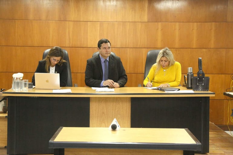 La jueza Natalia Cacavelos usa laptop, Darío Báez escucha atento y Gloria Hermosa toma notas, rodeados de documentos en ambiente formal.