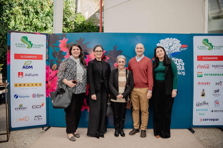 Cinco personas sonrientes posan de pie en un evento decorado con flores y logos de patrocinadores, en un ambiente festivo.