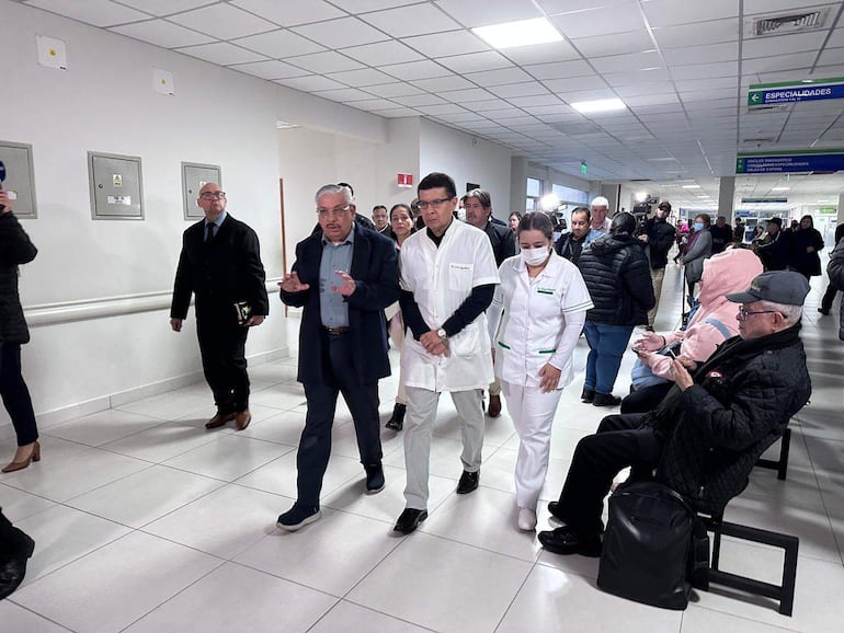 Isaías Fretes en camisa azul y abrigo oscuro, acompañado de personal médico en un pasillo del hospital, con pacientes en espera al fondo.