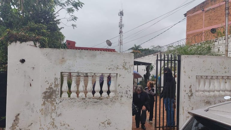 Mujer de cabello largo y gris se inclina junto a la puerta, acompañada por otra mujer con abrigo oscuro y varias personas con paraguas.