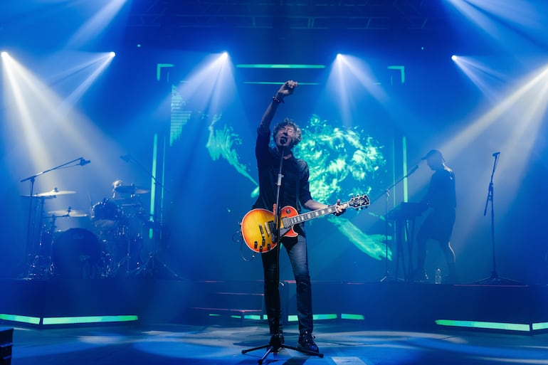 Guitarrista enérgico con guitarra naranja en un concierto, con luces brillantes y coloridas de fondo en el Gran Galpón.