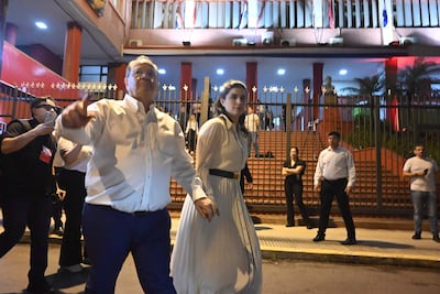 Camilo Pérez con camisa blanca y pantalones oscuros, junto a mujer en vestido blanco, conversando ante un grupo en evento exterior.