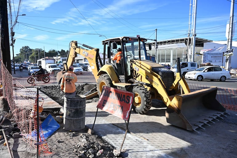 Dos trabajadores en ropa de alta visibilidad junto a una excavadora amarilla en sitio de construcción, rodeados de vehículos y vallas de seguridad.