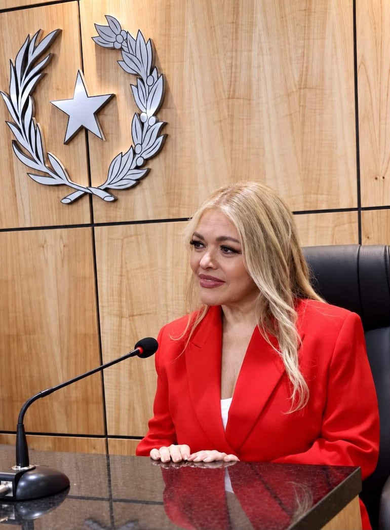 Mujer rubia de cabello largo y suelto en chaqueta roja, sentada frente a un micrófono en ambiente formal de madera.