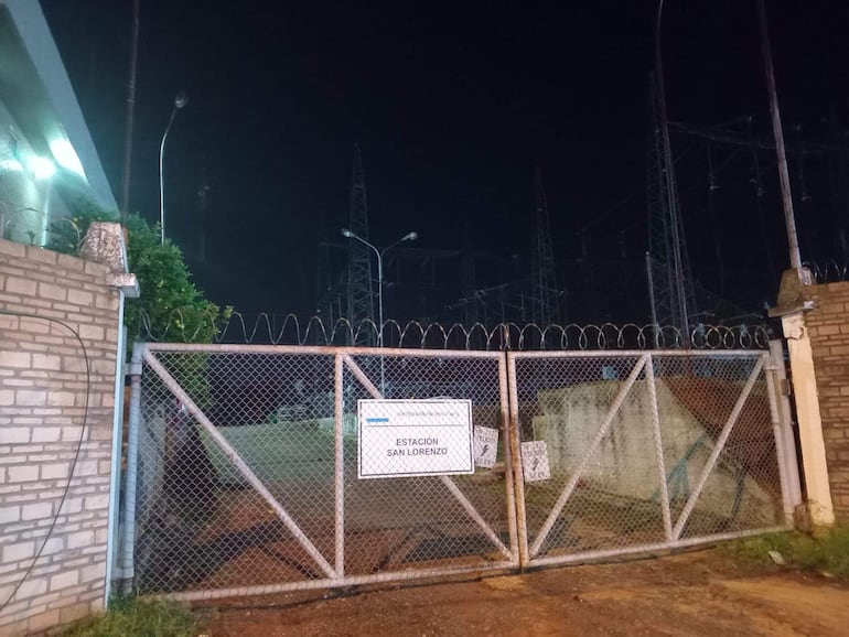 Entrada de la estación de la ANDE iluminada, con reja de seguridad y torres eléctricas al fondo en un cielo oscuro.