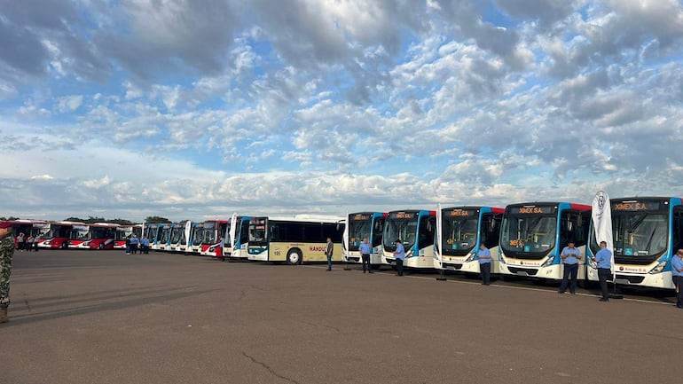 Empleados en uniformes azules rodean autobuses de colores brillantes, en un ambiente de espera bajo un cielo parcialmente nublado.