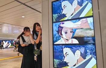 Dos jóvenes miran a una amiga mientras permanecen junto a una cartelera digital que anuncia el próximo lanzamiento del videojuego “Demon Slayer: Kimetsu no Yaiba – The Hinokami Chronicles 2”, a lo largo de un pasillo dentro de una estación de tren en el centro de Tokio, el 29 de julio de 2025.
