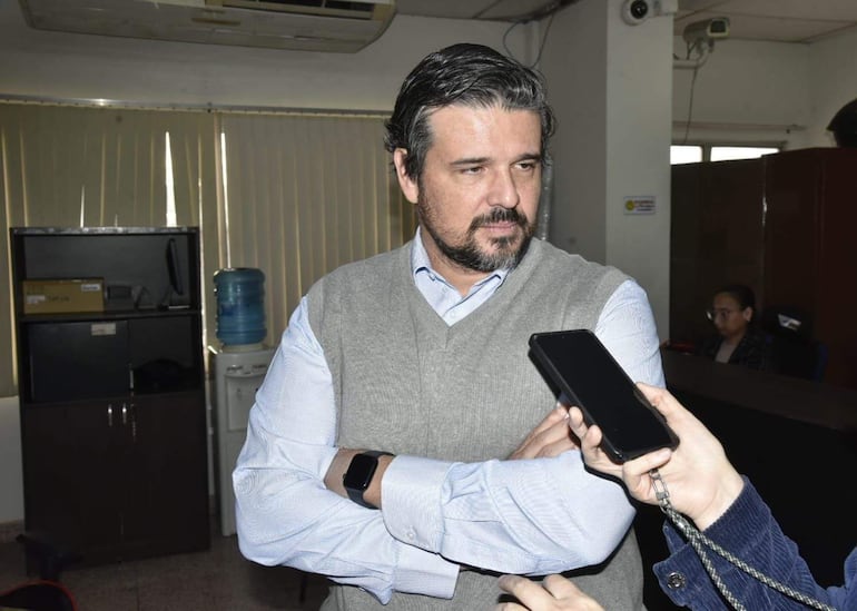 Juan Carlos Frutos, con suéter gris y camisa blanca, de pie en oficina, sostiene brazos cruzados mientras se le entrevista.
