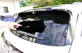 Persona con camisa blanca señala un gran agujero en la luneta trasera de un coche blanco en un estacionamiento.