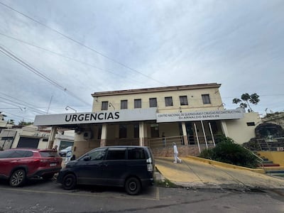 Hombre de pie frente a la edificación de dos pisos del centro médico, donde se observa un letrero de 'URGENCIAS'.
