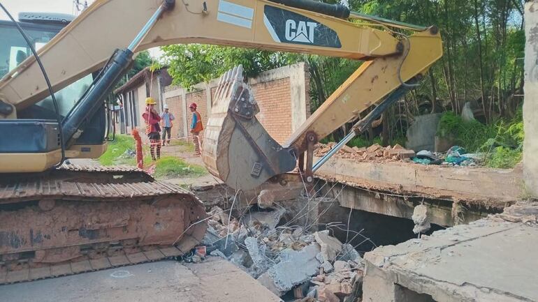 Cuatro trabajadores con cascos de seguridad y chaleco azul, revisan escombros mientras un excavador CAT levanta materiales en un entorno urbano.