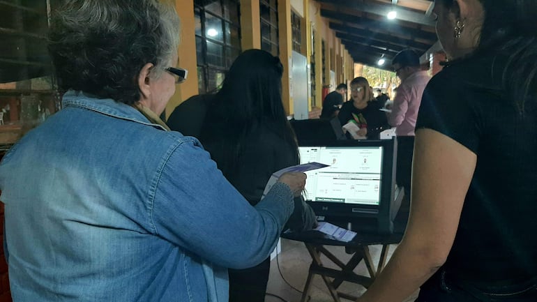 Mujer canosa en chaqueta de mezclilla sostiene documentos, observa pantalla; otra mujer atenta junto a ella en lugar de votación.