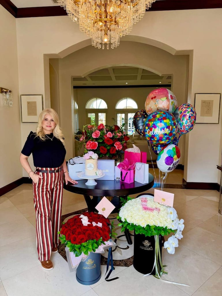 Mujer rubia en blusa negra y pantalones a rayas sonríe junto a mesa con tarta, globos y regalos coloridos.