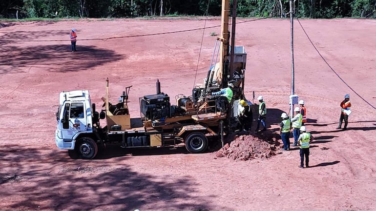 Grupo de trabajadores en terreno de construcción, con cascos blancos y chalecos de seguridad cerca de maquinaria de perforación.