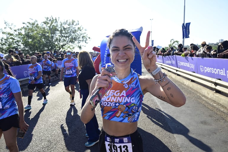 Competidora sonriente sostiene un trofeo y hace gesto de paz. Multitud y banners de 'MEGA RUNNING 2026' en el fondo.