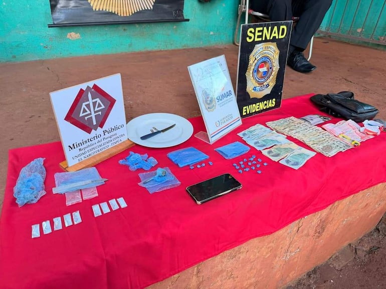 Mesa con envoltorios de drogas, billetes, un cuchillo y un teléfono móvil, todo bajo un mantel rojo, sin personas presentes.