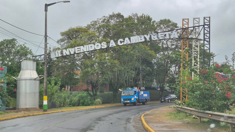 Camión azul en ruta con letrero de bienvenida a Cambyretá y estructura metálica de producción láctea al fondo.