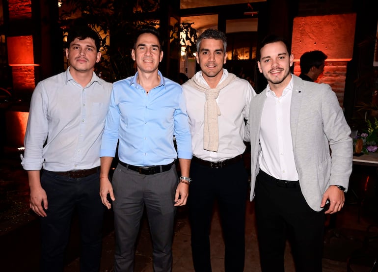 Cuatro hombres sonrientes en camisas y chaquetas, posan juntos en un ambiente iluminado con plantas como decoración.