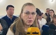 María Teresa Barán con gafas y abrigo negro, sostiene un micrófono frente a un grupo que la escucha atentamente en un ambiente serio.