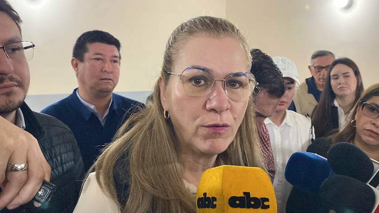 María Teresa Barán con gafas y abrigo negro, sostiene un micrófono frente a un grupo que la escucha atentamente en un ambiente serio.