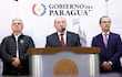 Hombre en traje oscuro y corbata roja habla en conferencia, flanqueado por dos hombres en ropa formal con expresiones serias.