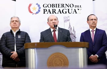 Hombre en traje oscuro y corbata roja habla en conferencia, flanqueado por dos hombres en ropa formal con expresiones serias.