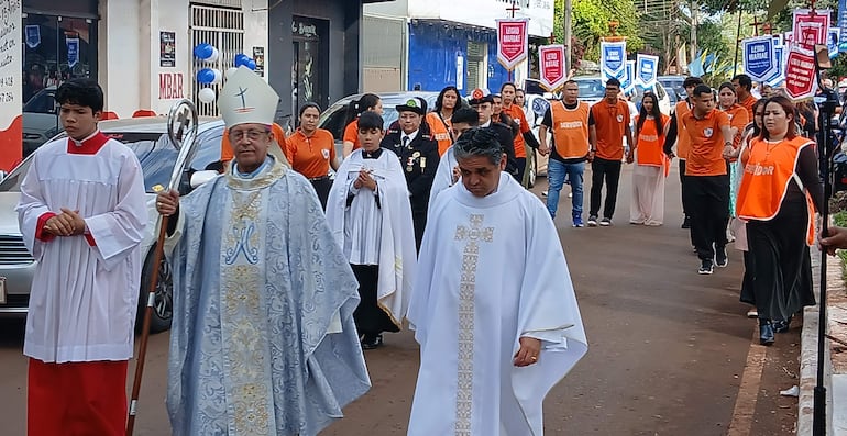 Sacerdote con báculo y monaguillo en procesión, rodeados de personas con camisetas naranjas y banderas en un ambiente urbano.