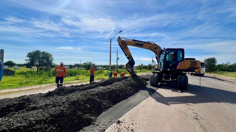 Cinco trabajadores en camisetas naranjas colaboran cerca de una excavadora en una zona rural con cielo despejado.