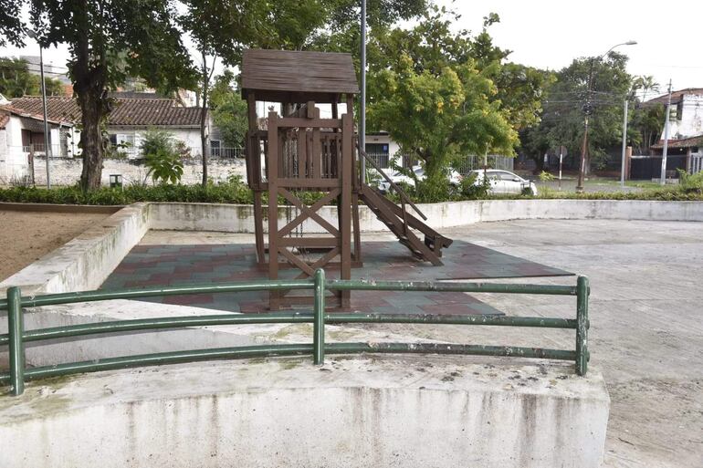 Área recreativa vacía con estructura de juegos de madera y baldosas de colores rodeada de plantas.