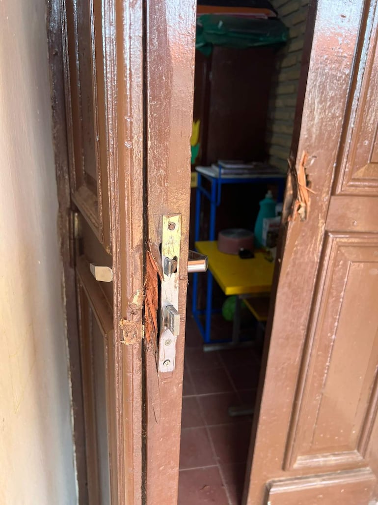 Puerta de madera de la escuela con daño visible, mostrando un aula con muebles amarillos y libros en el fondo.