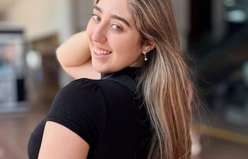 Mujer joven con cabello largo y lacio, sonríe vistiendo camiseta negra en un ambiente público.