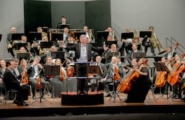 Luis Szarán, con chaqueta y corbata, dirige a una orquesta de 40 músicos en un elegante teatro.