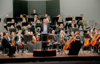 Luis Szarán, con chaqueta y corbata, dirige a una orquesta de 40 músicos en un elegante teatro.