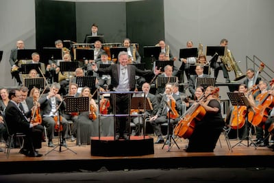 Luis Szarán, con chaqueta y corbata, dirige a una orquesta de 40 músicos en un elegante teatro.